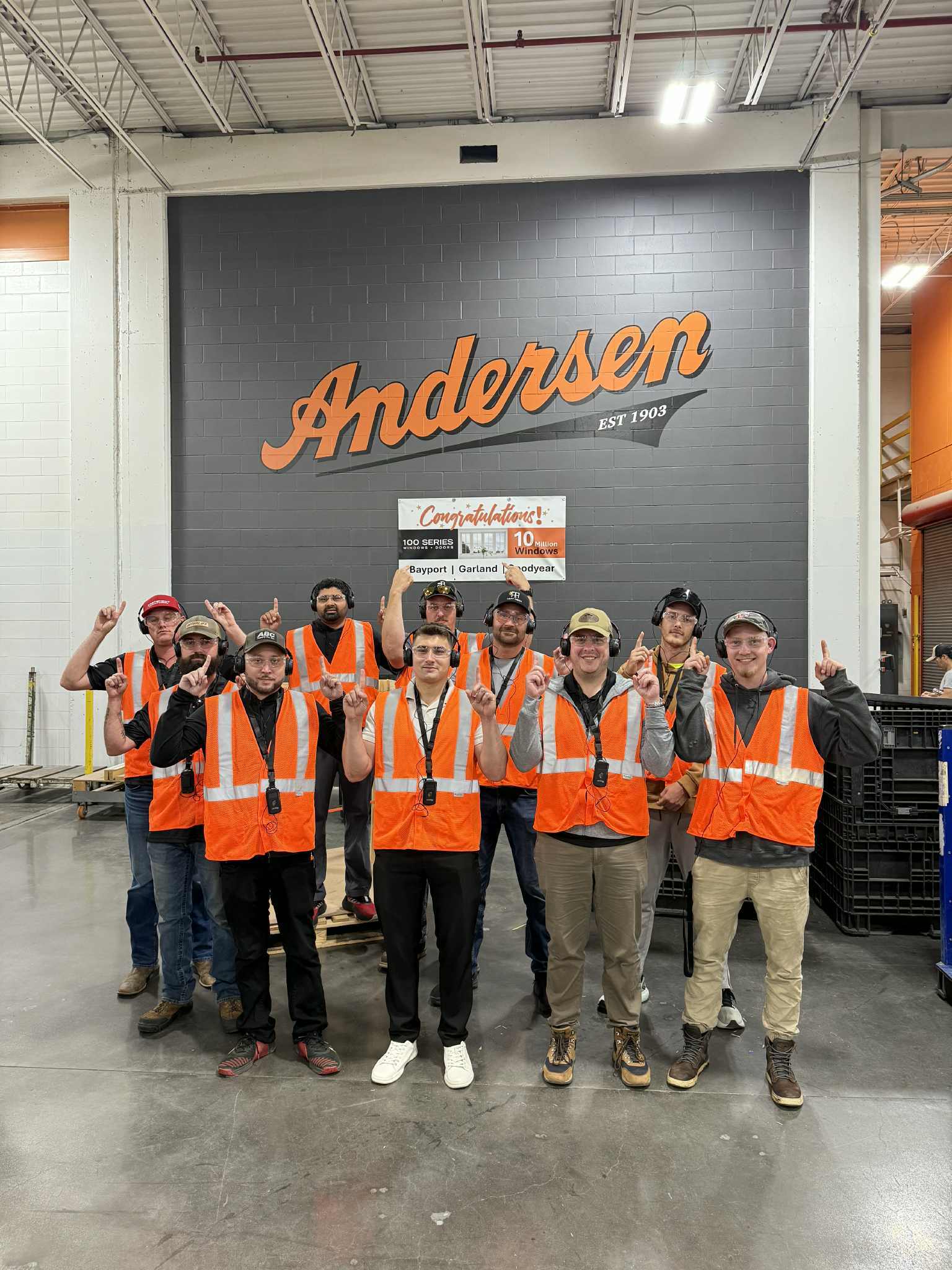 A group of workers wearing orange safety vests and hats pose together indoors in front of a gray wall with the Andersen logo, showcasing team spirit and dedication. Learn more on our About Us page.