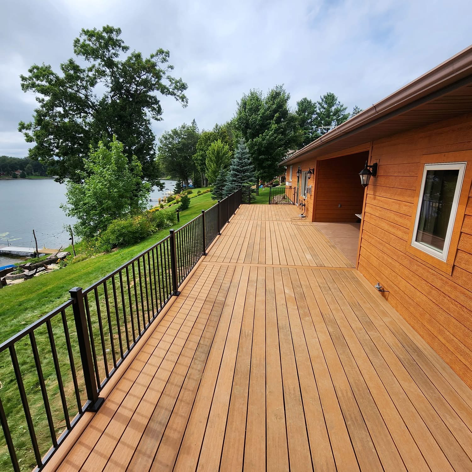 Spacious wooden deck with black metal railing alongside a house, overlooking a grassy yard and a lake with trees in the background.