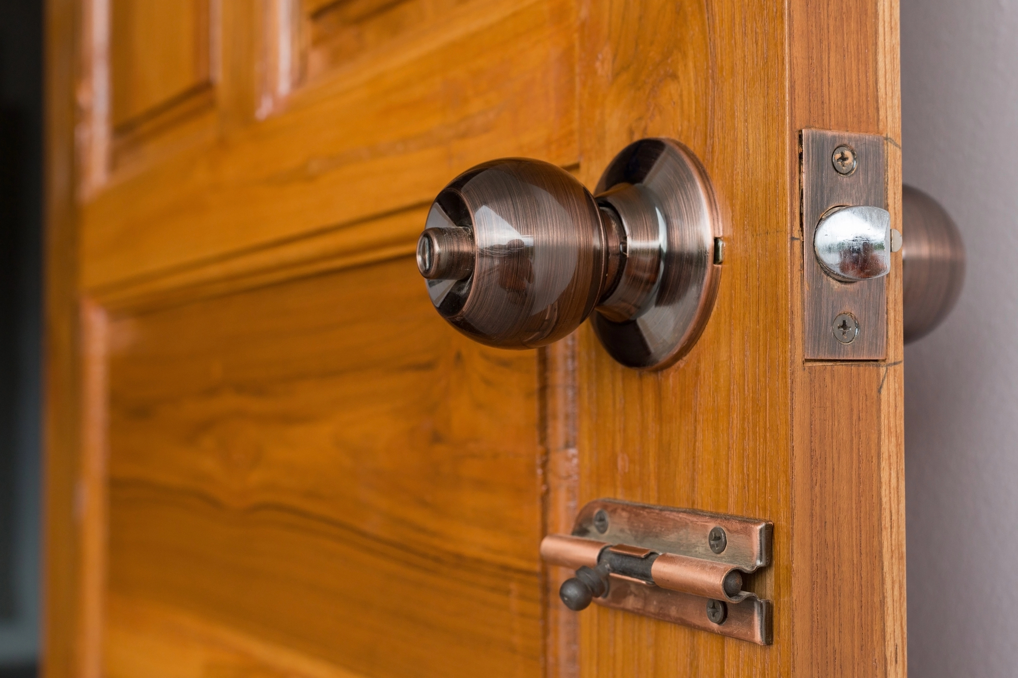 A wooden door slightly open, showing a round metallic door knob and a sliding bolt latch installed below the knob.