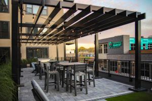 Outdoor rooftop patio with gray tables and chairs under a black pergola with string lights, overlooking nearby buildings including a market.