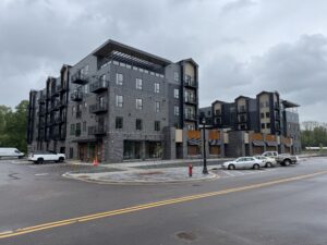 A modern five-story apartment building with balconies, adjacent parking lot, and cars parked in front on a cloudy day.
