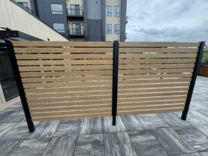 A horizontal slat privacy fence with wood panels and black metal posts stands on a tiled patio in front of a modern apartment building.