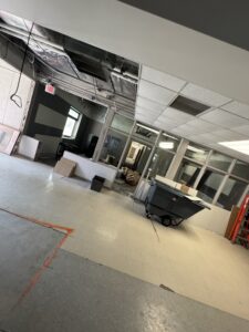 A partially renovated office space with missing ceiling tiles, exposed ducts, scattered debris, and a cleaning cart near glass-walled rooms.