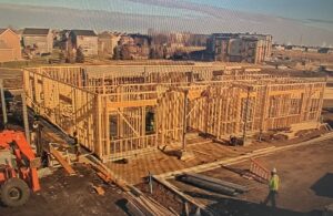 A building under construction with a wooden frame structure, construction equipment, workers, and nearby residential buildings in the background.