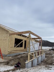 A partially constructed wooden building stands on a snowy lot, with exposed framing, ladders, and construction materials visible.