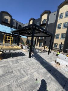 Outdoor construction site with a partially built pergola, tools, materials, and unfinished brickwork on a paved surface in front of a modern apartment building.