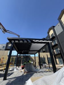 A black metal pergola under construction in a courtyard, surrounded by tools, materials, and adjacent apartment buildings under clear blue sky.