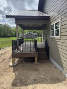 A covered wooden porch with black railings and stairs attached to a tan house, overlooking a grassy yard and distant trees.