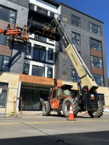 Construction workers use a lift and a telescopic handler to work on the exterior of a multi-story building, with traffic cones marking off the area.