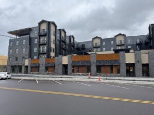 A modern four-story apartment building with ground-floor commercial spaces, empty parking lot, and overcast sky.