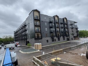 A newly constructed four-story apartment building with balconies, surrounded by a wet parking lot and construction materials on a cloudy day.