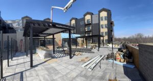Outdoor construction site with metal pergolas being built on a tiled patio, surrounded by tools, materials, and multi-story apartment buildings in the background.