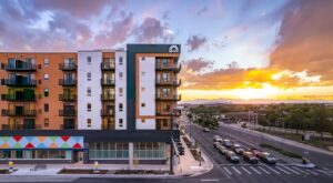 Modern mid-rise apartment building with balconies at sunset, located on a busy street with multiple cars and a view of distant mountains.
