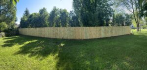 A newly installed wooden privacy fence runs along the edge of a grassy backyard, with tall green trees in the background.