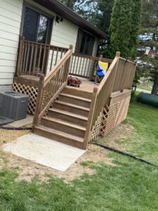 A wooden deck with railings and steps leads up to a house. A lawn chair and leaf blower are on the deck; an air conditioning unit is next to the stairs.