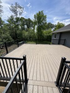 A spacious wooden deck with black metal railings is attached to a gray house, overlooking a grassy yard and trees under a partly cloudy sky.