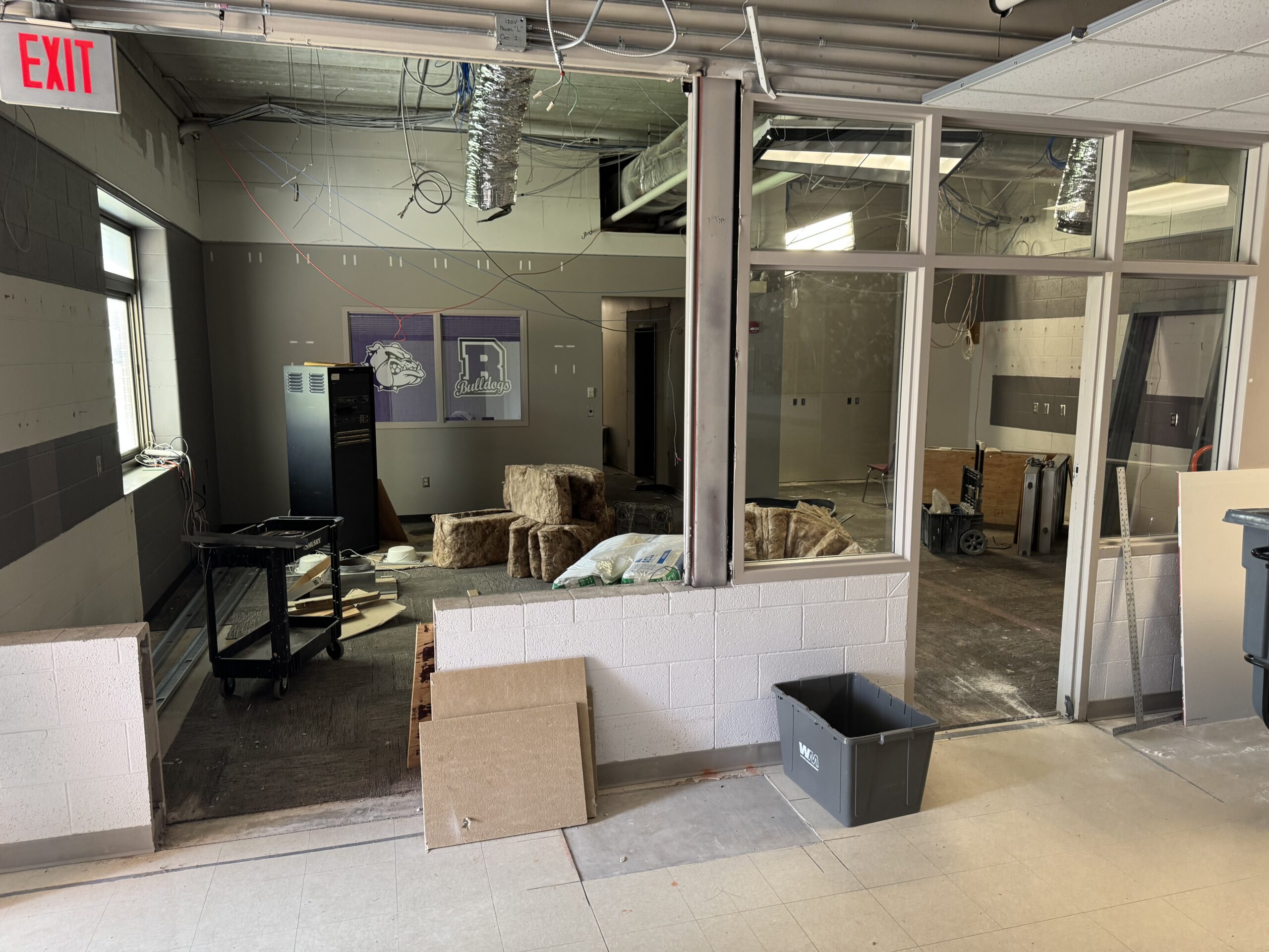 A partially renovated room undergoing remodeling, with construction materials, exposed ducts, a bin, and furniture stacked in the corner; windows separate the room from the hallway.