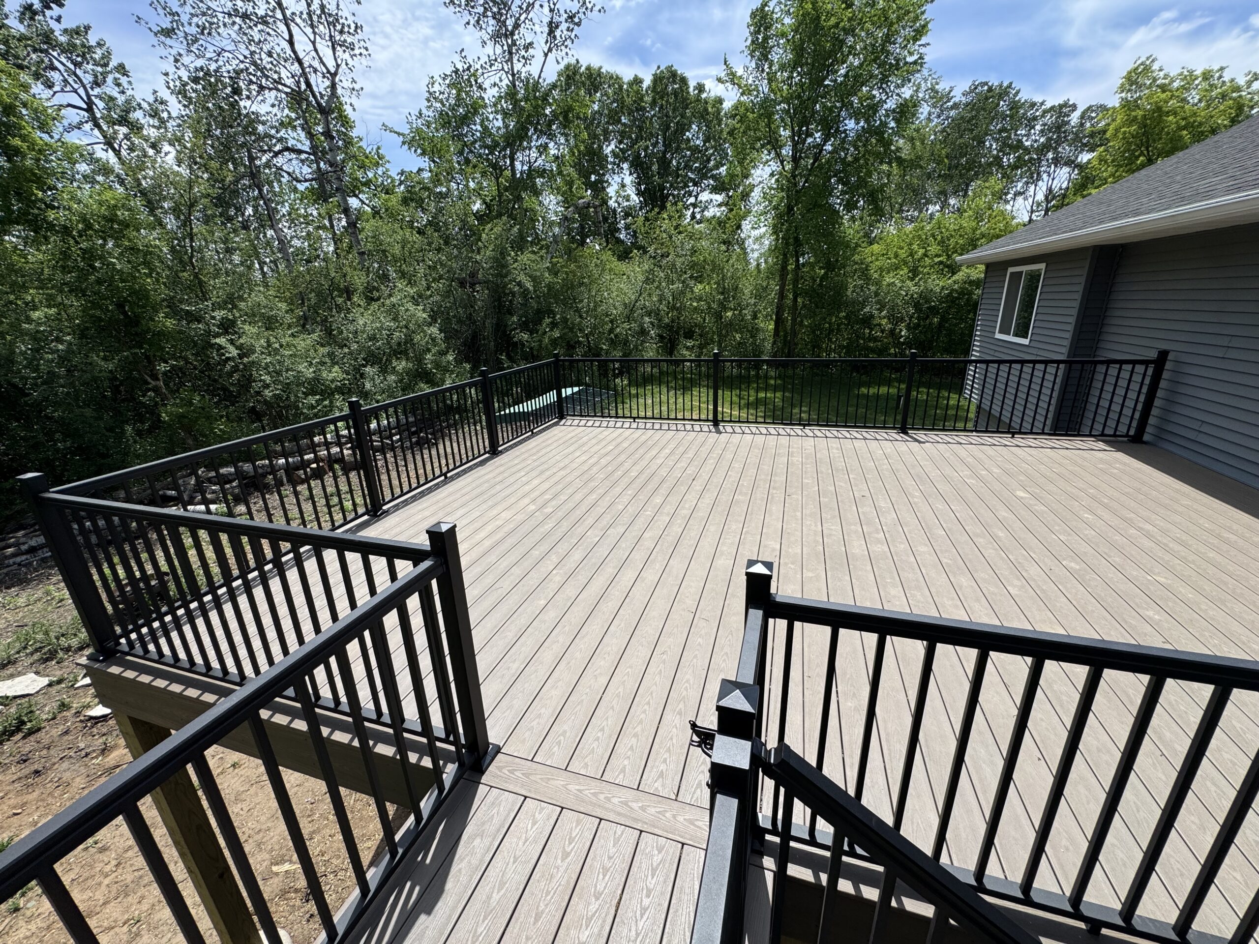 A spacious, elevated wooden deck with black metal railings overlooks a grassy yard with trees beside a gray house, perfect for relaxing after a window installation service.