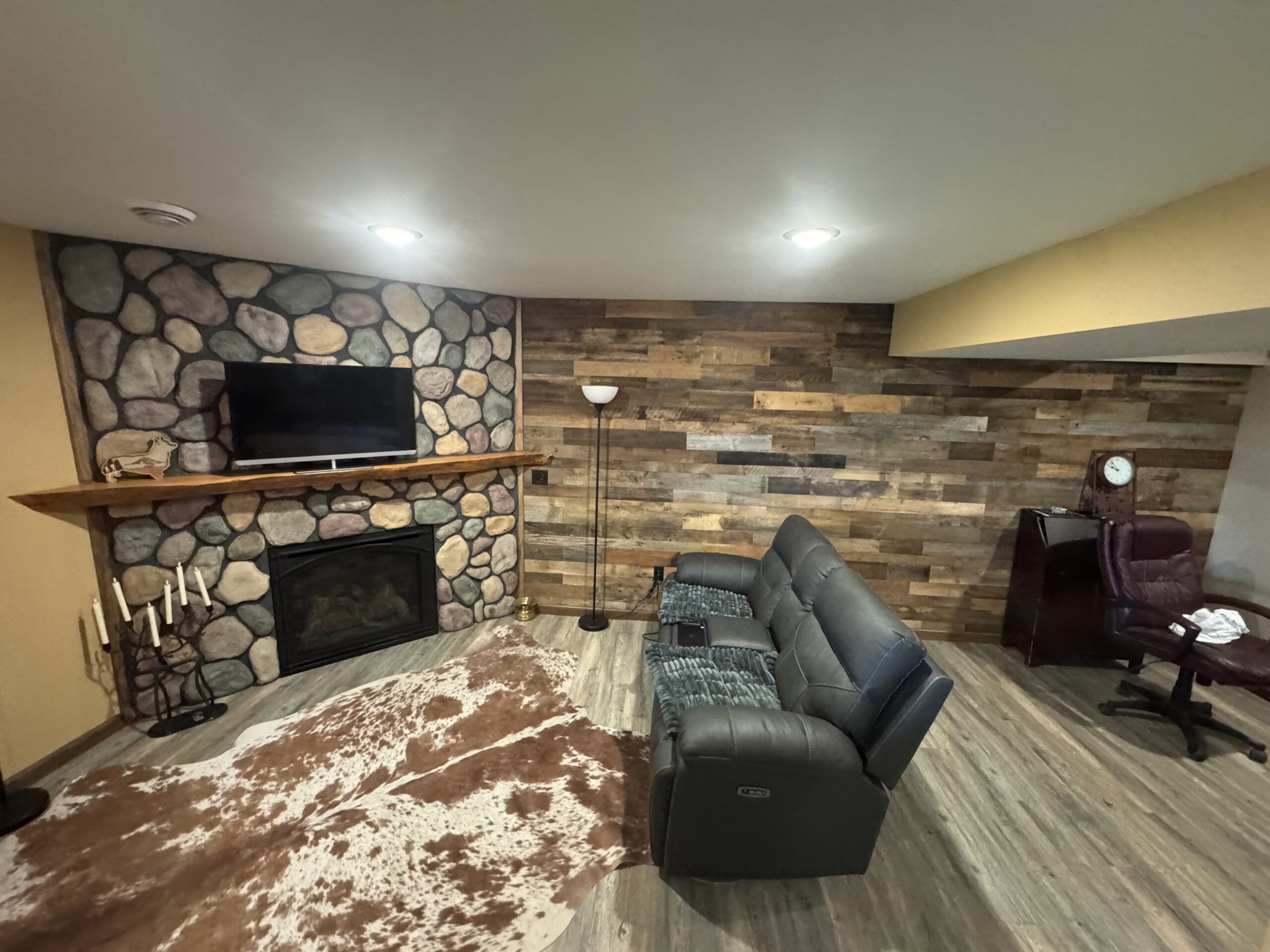 Basement living room with stone fireplace, mounted TV, cowhide rug, leather reclining sofa, standing lamp, wood-paneled wall, and a desk with a chair in the corner—perfect for relaxing or considering a window installation service.