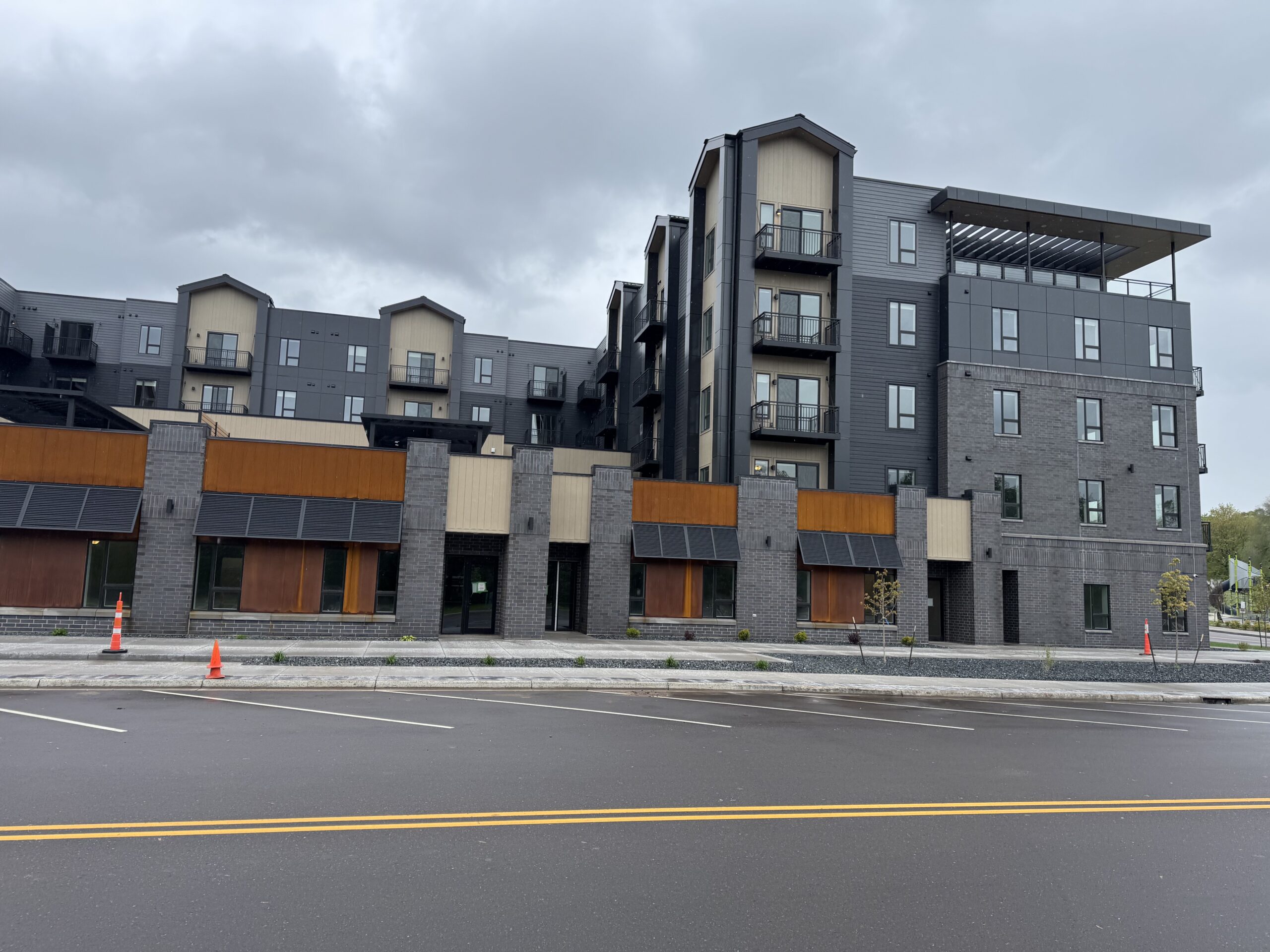 A modern multi-story apartment building with closed ground-floor retail spaces, empty sidewalks, and overcast skies.