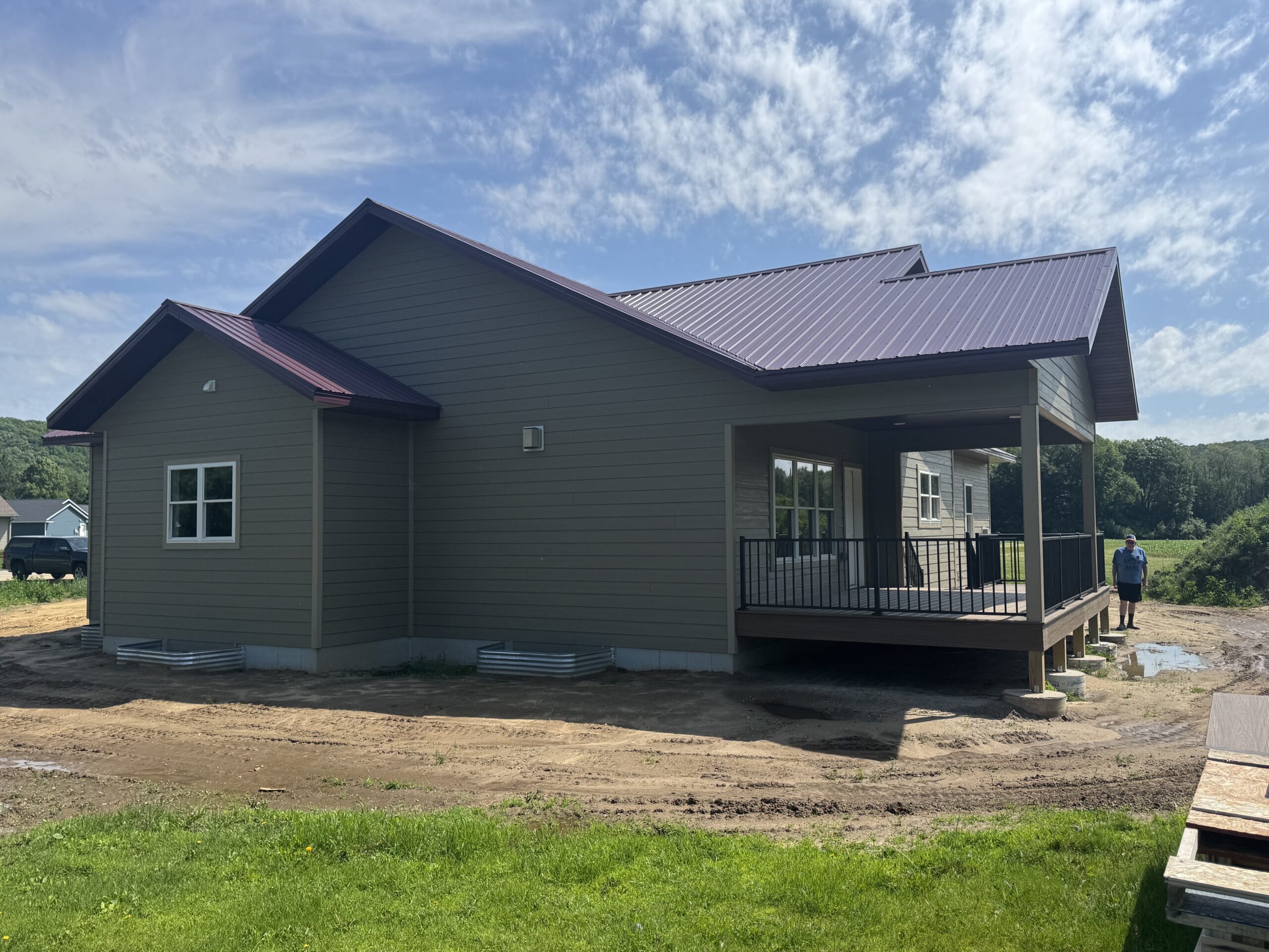 Single-story house with gray siding and a metal roof, featuring a covered porch; a person stands near the back, and the area around the house is unlandscaped. Durable siding enhances the home's clean, modern look.