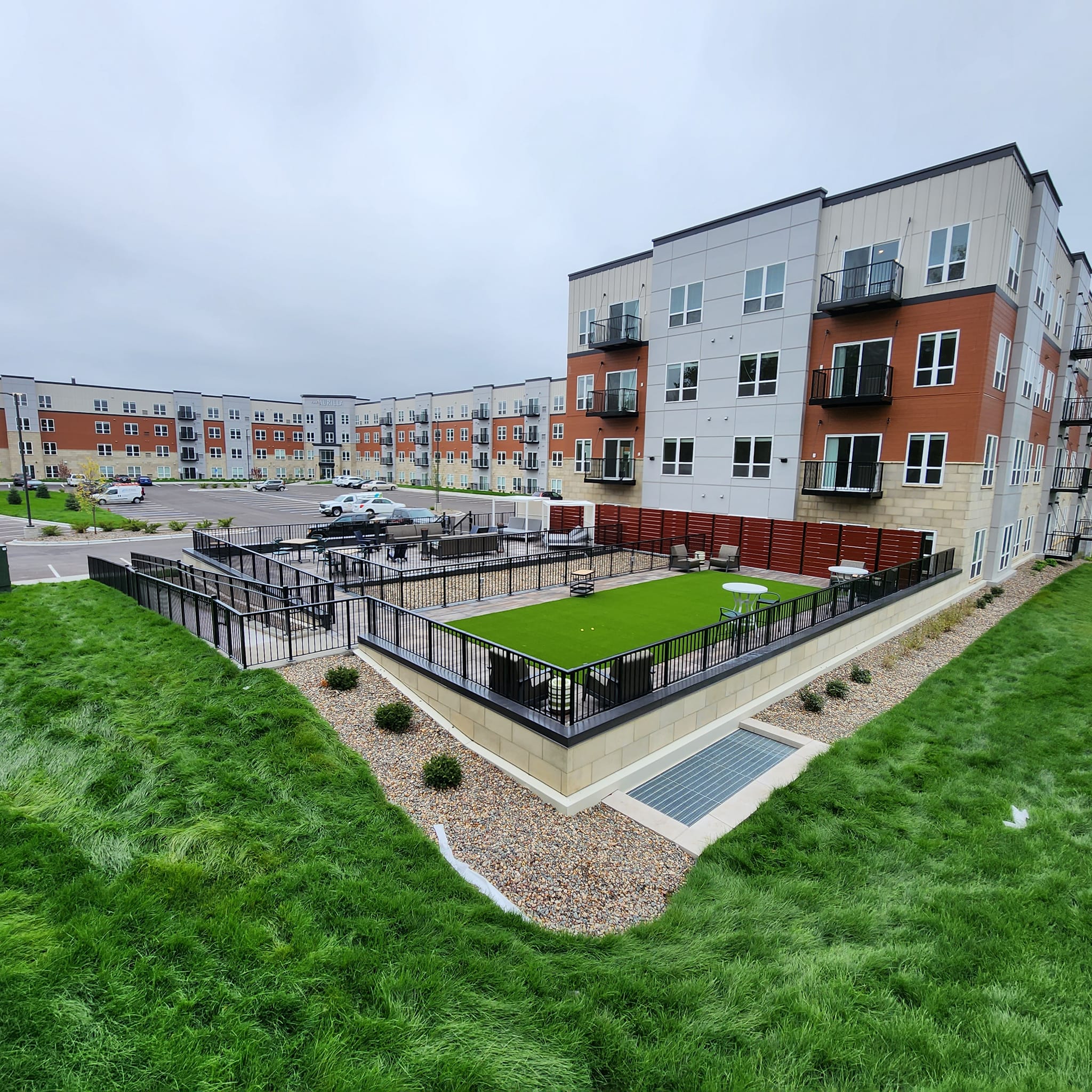 A modern apartment complex with a fenced outdoor area featuring artificial turf, tables, and chairs, surrounded by green grass and parking spaces.
