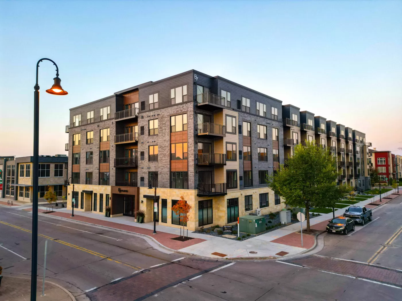 A modern four-story apartment building with balconies stands at the corner of a quiet intersection, surrounded by sidewalks, trees, and streetlights at dusk.