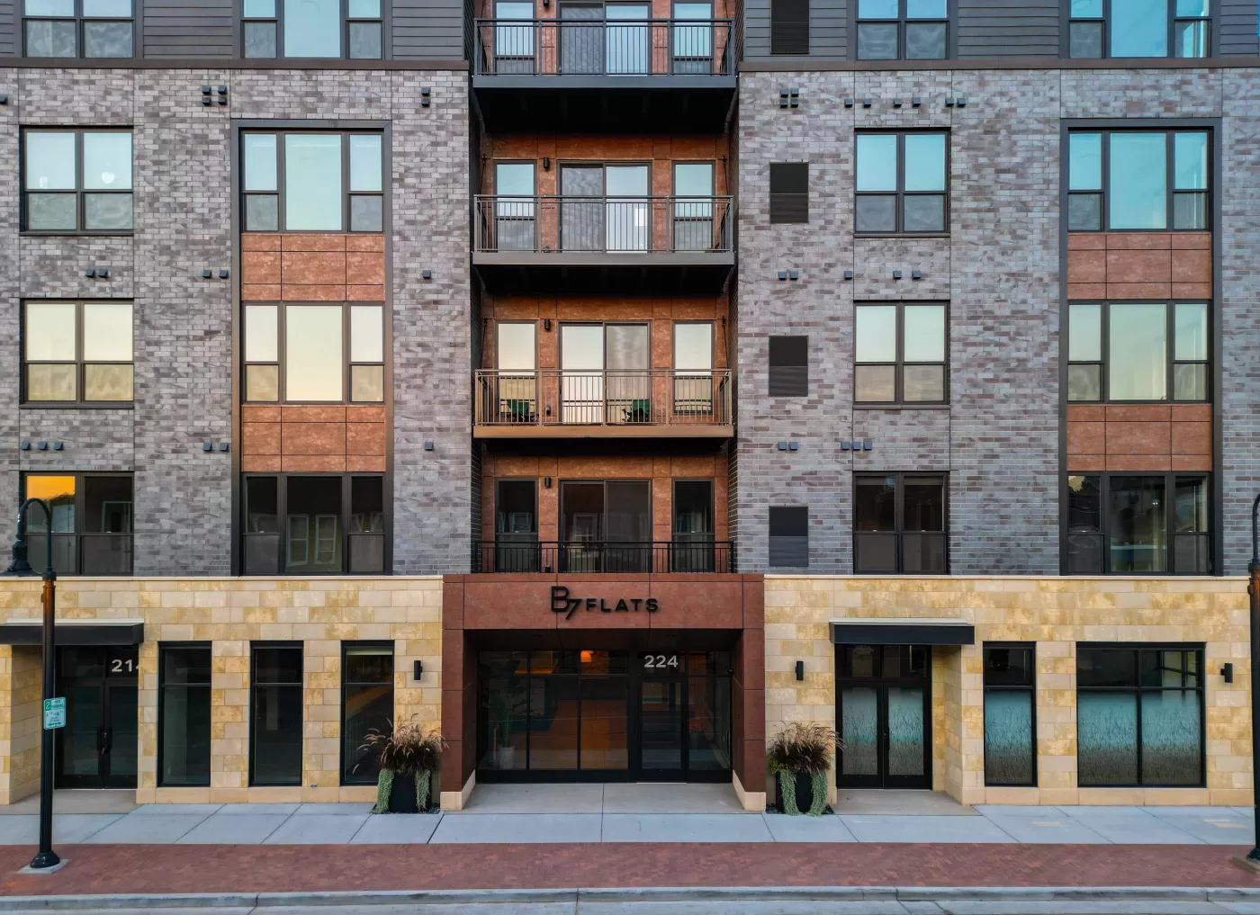 Modern apartment building exterior featuring large windows, a blend of brick, wood, and stone finishes, stylish balconies, and an entrance labeled B Flats above sleek glass doors.